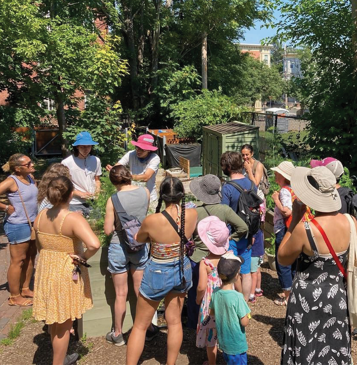 A group of people gathered in an outdoor garden setting, some wearing sunhats. Trees and garden structures are visible in the background.