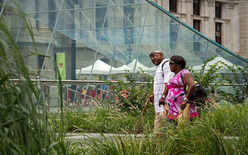 A couple walks between too tall grass plantings leading to the center of the plaza