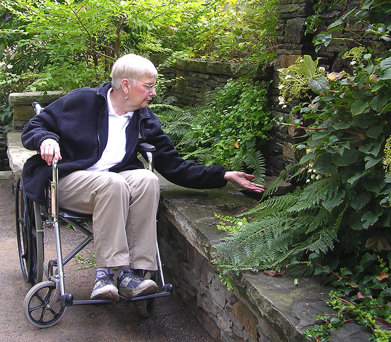 A women in a wheelchair reaches over a low wall to touch a water feature located just off the path of the garden.