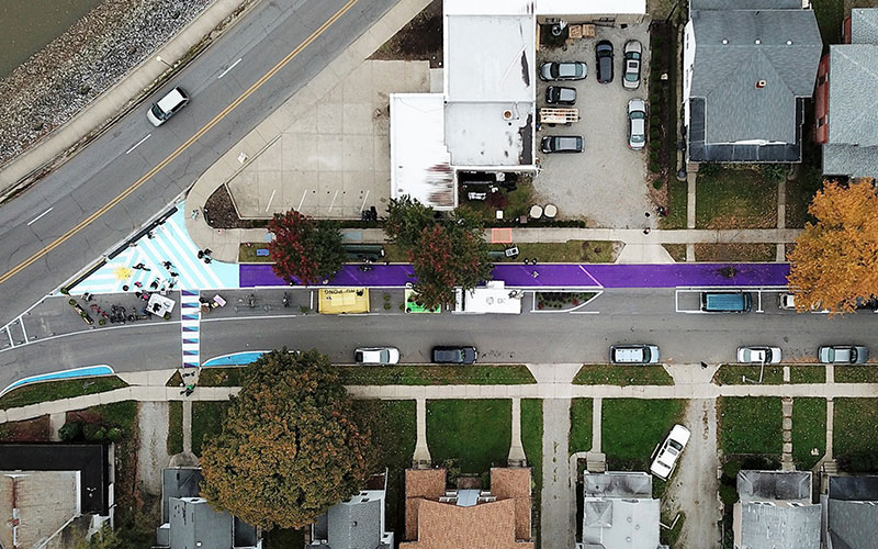 Aerial view of the a bike path indicated by purple street paint, a series of small parklets, and a small plaza at the street corner.